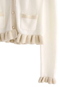 White wool cardigan with beige ruffled cuffs and hem. Features two front pockets with subtle texture and round buttons. Soft and cozy design.