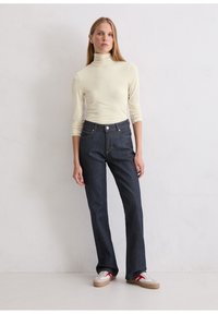 Cremefarbener Rollkragenpullover, kombiniert mit dunkelblauen geraden Jeans, die über fünf Taschen, einen Knopfverschluss und subtile orangefarbene Stickdetails verfügen.