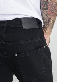 Jeans de mezclilla negra con un acabado texturizado. Las características incluyen una etiqueta de cuero con letras en relieve y costuras visibles en los bolsillos traseros.