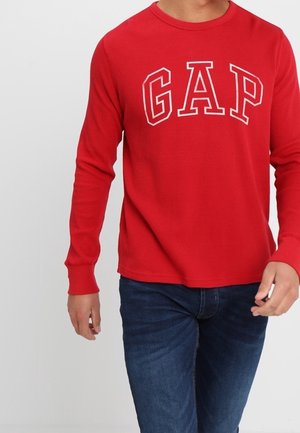 Rød langærmet skjorte med hvide konturerede "GAP" bogstaver på tværs af brystet båret med mørkeblå jeans på en person.