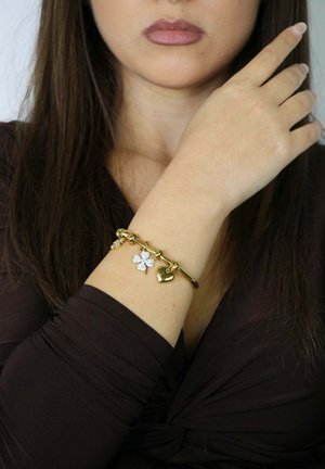 Frau in dunklem Oberteil zeigt ein goldenes Armband mit Herz- und Kleeblattanhängern am erhobenen Handgelenk nahe am Kinn.