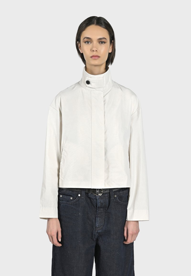 Giacca bianca Crop con colletto alto, chiusura frontale con bottoni a pressione e tasche laterali; abbinata a pantaloni in denim blu scuro. Texture liscia.
