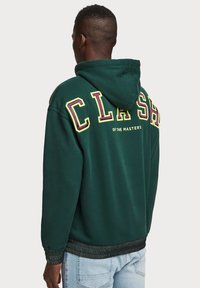 Sudadera verde con capucha ajustable, con la palabra "CLASH" en letras amarillas y moradas en la parte posterior, y detalles de ribete negro en el dobladillo.