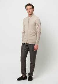 Suéter beige de cremallera en tela suave, diseño ajustado con cuello alto. Combinado con pantalones de lana gris y zapatos de ante oscuros.