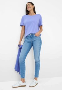 T-shirt léger violet à manches courtes associé à un jean bleu serré et des baskets blanches. Le mannequin tient un pull violet assorti.