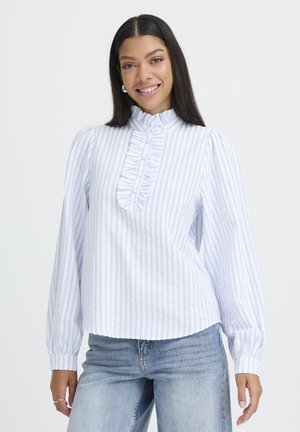 Lächelnde Frau mit langen dunklen Haaren, die eine weiße Bluse mit hellblauen Streifen, Rüschendetail und hellblauen Jeans vor einem schlichten Hintergrund trägt.