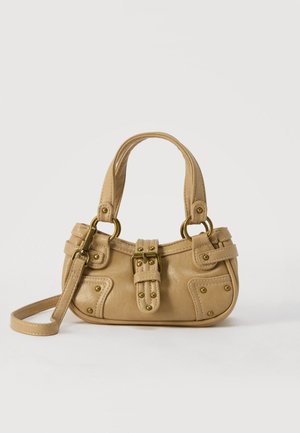 Sac à main en cuir beige avec double poignées, boucle en laiton et détails rivetés, ainsi qu'une bandoulière réglable sur fond blanc.