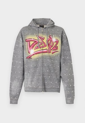 Young man stands wearing a grey hoodie with red graffiti text, faded blue jeans with a patch on the thigh, and green lace-up shoes.