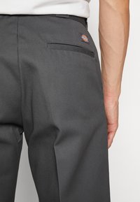 Grijze broek met een gladde textuur, voorzien van een achterzak en een logo-label op de tailleband. Close-up van opzij.