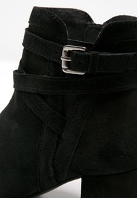 Bottine en daim noir avec une sangle autour de la cheville et une boucle argentée sur un talon carré.