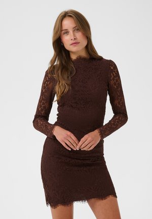 Femme aux longs cheveux châtain clair porte une robe moulante en dentelle marron foncé à manches longues, debout devant un fond clair uni.