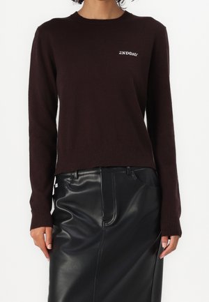 Personne portant un pull marron foncé avec le texte "2NDDAY" et une jupe en cuir noire, debout devant un fond blanc uni.