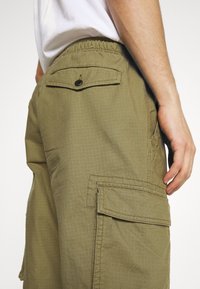 Pantalon cargo vert olive avec une finition texturée, taille élastique, poches latérales et une poche arrière avec fermeture par bouton noir.