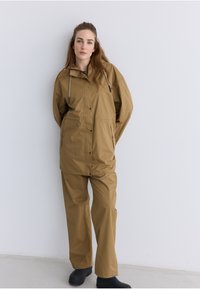 Hellbraune wasserdichte Jacke mit Kapuze und Zugbändern, kombiniert mit passenden Hosen. Das Material wirkt glatt und leicht glänzend. Schwarze Fußbekleidung.