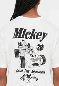 Bílý tričko s černou grafikou Mickey Mouse v závodním autě, s texty "Mickey," "28," a "Grand Prix Adventures". Součástí jsou také kostkované vlajky.