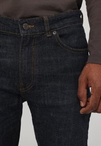 Ciemnoniebieskie dżinsy z pomarańczowymi stębnowaniami, pięciopocketowy fason, metalowe zapięcie i teksturowane wykończenie. Zbliżenie na talię i rękę.