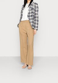 Blazer a scacchi blu e bianco, pantaloni beige con vita elastica e sandali leggeri con sottili cinturini. Texture morbide e vestibilità sartoriale.