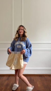 Žena stojící uvnitř na dřevěné podlaze, oblečená v modrém oversized svetru, bílých šortkách a pantoflích, držící žluté šortky před sebou.