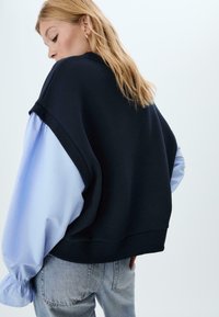 Felpa oversize di un blu scuro con maniche ampie di un blu chiaro. Polsini e orlo a costine, con una vestibilità comoda. Parte dei pantaloni in denim visibili.