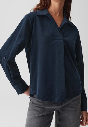 Kvinde iført en mørkeblå skjorte med lange ærmer, knaplukning ved manchetterne og grå jeans, stående mod en ensfarvet lys baggrund.