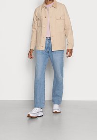 Beige bomulljacka med två framfickor, knappstängning och en lätt design, kombinerad med ljusblå jeans och vita sneakers.