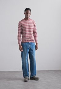 Rood-wit gestreepte gebreide trui met lange mouwen, gecombineerd met losse blauwe denim jeans en grijze schoenen, staand tegen een neutrale achtergrond.