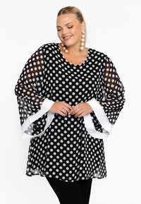 Transparente schwarze Bluse mit weißen Punktmotiven, Glockenärmeln und weißen Besätzen, die mit kleinen Pompons verziert sind. Lockere Passform, runder Ausschnitt.