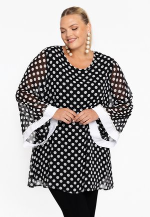 Doorzichtig zwart blouse met witte stippen, klokmouwen en witte bies versierd met kleine pomponnetjes. Losvallend, ronde halslijn.