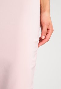 Vaaleanpunainen säädettävä mekko, jossa on sileä pinta, saumatonta muotoilua ja polveen ulottuva leikkaus, jossa käsi pitää hellästi kangasta.