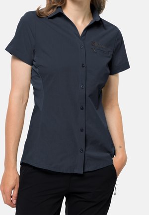 Femme portant une chemise bleu marine foncé cintrée à manches courtes avec boutons, une poche poitrine zippée et un logo sur le côté gauche.
