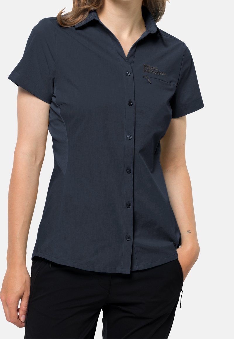 Mujer que lleva una camisa azul marino oscuro ajustada, de manga corta, con botones, con un bolsillo en el pecho con cremallera y logo en el lado izquierdo.