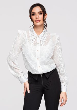 Femme en blouse blanche en dentelle transparente avec col et poignets ornés, associée à un pantalon noir taille haute, posant devant un fond uni.