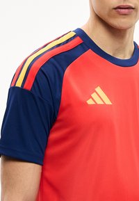 Άνδρας που φοράει κόκκινη και ναυτική αθλητική μπλούζα με κίτρινες και κόκκινες ρίγες στον ώμο και κίτρινο λογότυπο στο στήθος, περικομμένος στο πηγούνι και το πάνω μέρος του μπράτσου.