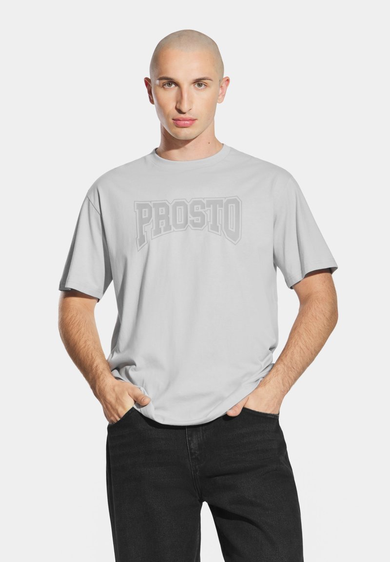 Jasnoszara koszulka z krótkim rękawem z odważnym, teksturowanym logo "PROSTO" na piersi, noszona z czarnymi dżinsami.