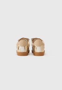 Sneaker beige con tacco metallico dorato. Presentano una forma arrotondata, una tomaia liscia e dettagli testurizzati. La suola in gomma offre aderenza.