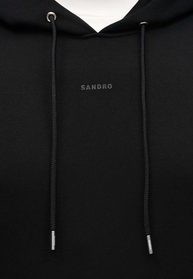 Schwarzer Hoodie mit kleinem grauem "SANDRO"-Logo auf der Brust, ausgestattet mit schwarzen Kordeln mit silbernen Metallenden und weißem Stoff, der im Inneren der Kapuze sichtbar ist.