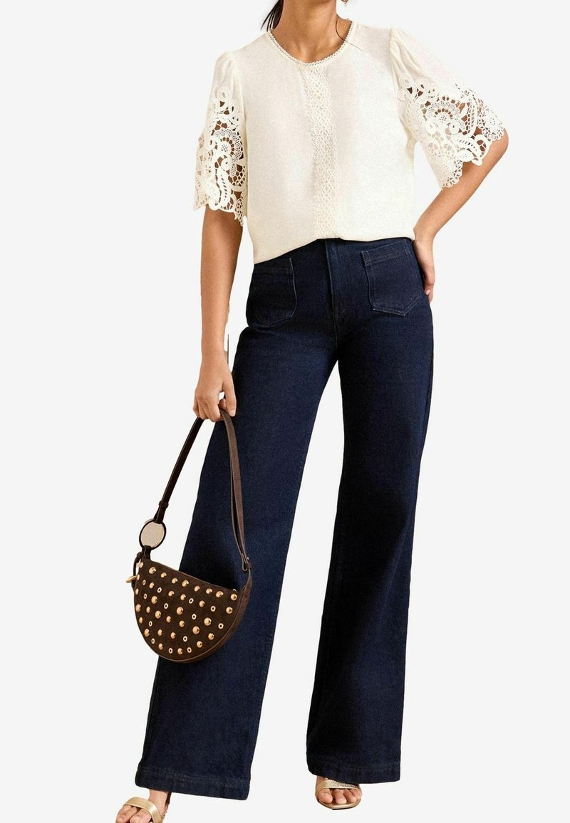 Femme portant un haut à manches en dentelle blanche et un jean large taille haute foncé, tenant un petit sac à main marron clouté d'une main.