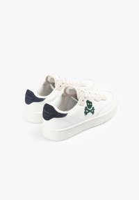 Zapatillas bajas blancas con acentos en el talón color marino, área de cordones beige y emblema de calavera y tibias cruzadas verdes en los laterales, mostradas desde ángulos trasero y lateral.