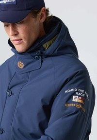 Marine vandtæt jakke med høj krave, trykknapper og broderede logoer. Har kontrastfarvet indre stof og tekstdetaljer på ærmet.