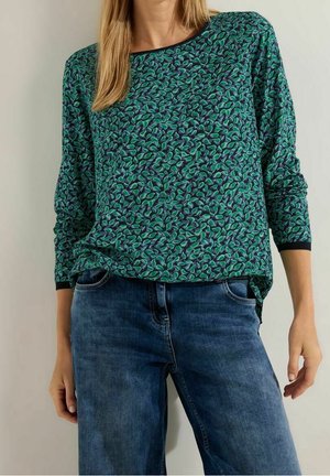 Frau trägt ein langärmeliges grün-blau gemustertes Oberteil mit dunkelblauen Jeans, steht mit entspannten Händen an den Seiten.
