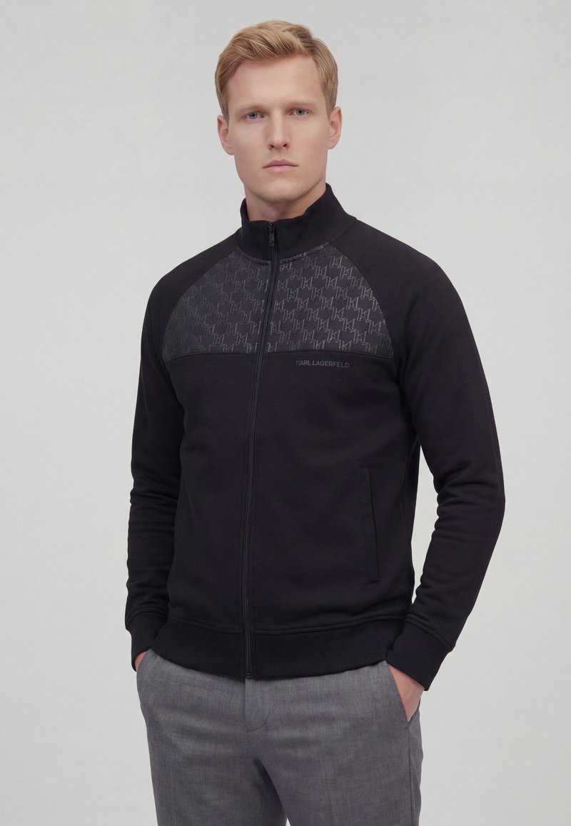 Junger Mann trägt eine schwarze Reißverschlussjacke mit gemustertem Oberteil und graue Hose, steht vor einem schlichten hellen Hintergrund.