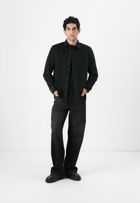 Dark green corduroy shirt with a front pocket, paired with black wide-leg jeans and black shoes, against a white background.