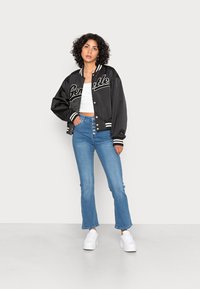 Casaco bomber em satin preto com letras brancas, acabamentos em malha riscada, combinado com jeans flare de tom azul claro e ténis brancos.