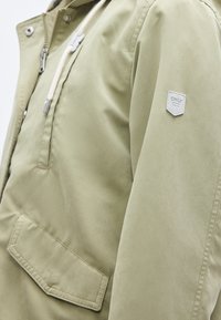 Ανοιχτό πράσινο μπουφάν από απαλό ύφασμα, με φερμουάρ, ρυθμιζόμενα κορδόνια και ετικέτα-μπάλωμα στο αριστερό μανίκι.