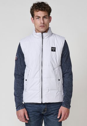 Chaleco acolchado blanco con cuello alto, cierre de cremallera y bolsillos frontales. Camisa de manga larga azul visible debajo. Pantalones de denim casuales.