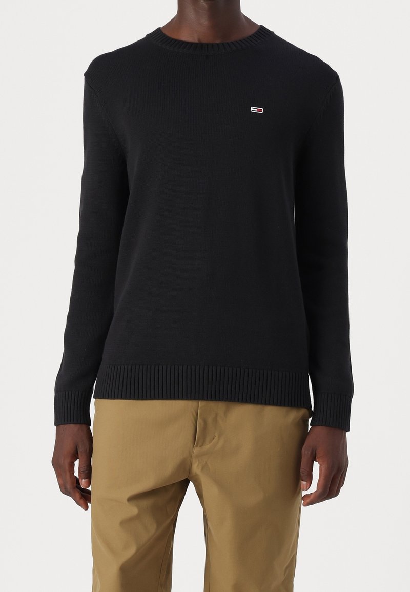 Homme portant un pull ras du cou noir avec un petit logo sur la poitrine et un pantalon beige, debout devant un fond uni.