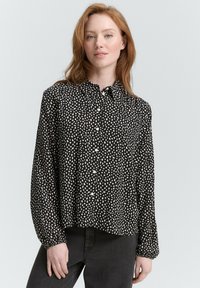 Chemise noire à boutons avec un motif abstrait de points blancs, manches longues et ourlet arrondi. Fabriquée en tissu léger avec une coupe décontractée.