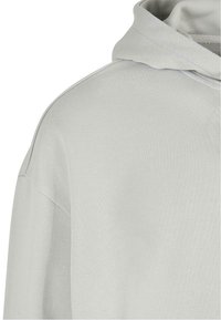 Sudadera gris claro hecha de una tela suave y texturizada. Presenta una capucha, hombros caídos y detalles de costura simples sin adornos.