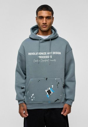 Capuchontrui in zacht teal stof, met de tekst "REVOLUTIONIZE ART DESIGN" in het wit. Voorzakt en verfspetters in blauw en zwart.