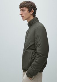 Giacca bomber imbottita verde scuro con colletto alto, tasche laterali e polsini a costine. Tessuto liscio con dettagli di design minimi.